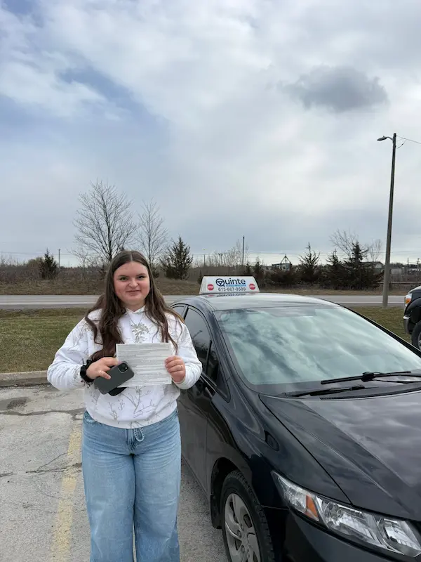 Young woman wearing a white top holding her road test passing paper beside a Quinte Driving School car.
