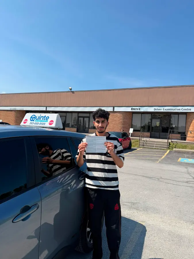 Young man wearing a striped t-shirt holding his road test passing paper beside a Quinte Driving School car.