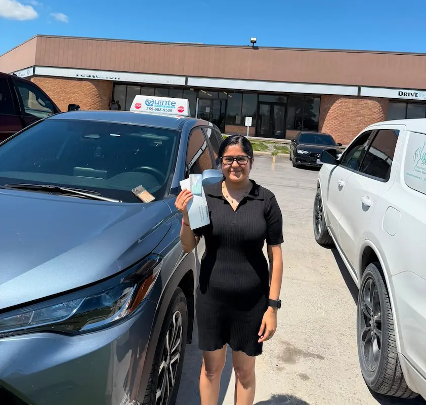 Smiling woman wearing a black dress holding her road test passing paper and her license while standing beside a Quinte Driving School car.
