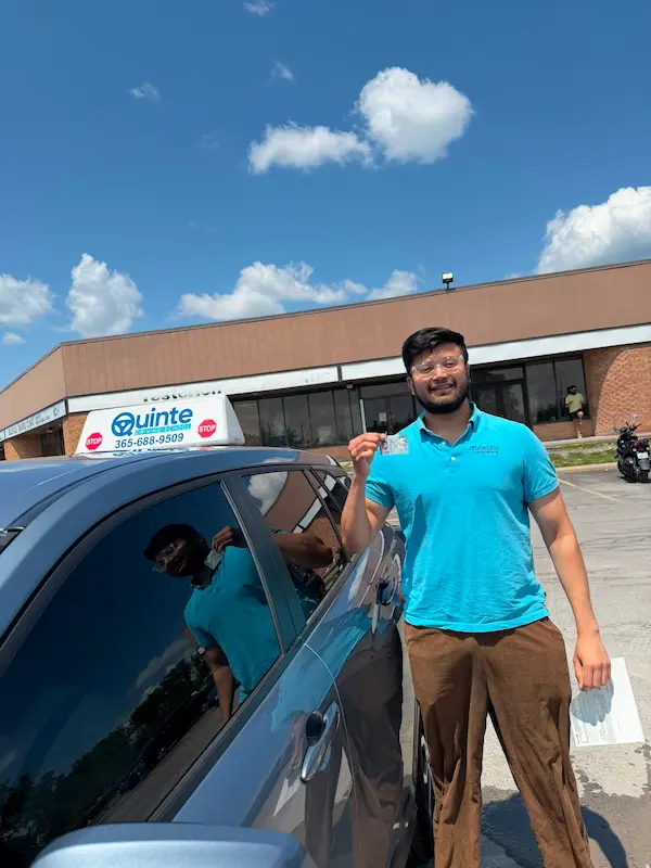 Smiling man wearing a blue polo shirt holding his road test passing paper and his driving license beside a Quinte Driving School car.