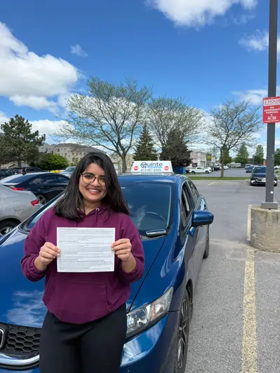 Smiling woman wearing a purple hoodie holding her road test passing paper in front of a Quinte Driving School car.