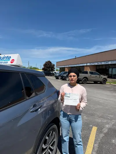 Smiling woman wearing glasses and a turban holding her road test passing paper beside a Quinte Driving School car.