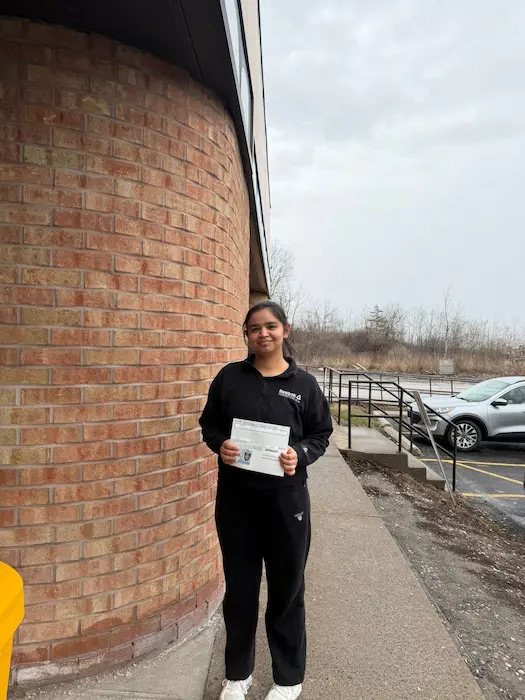 Smiling woman wearing a black outfit holding her road test passing paper and her driving license standing outside a driving test centre.