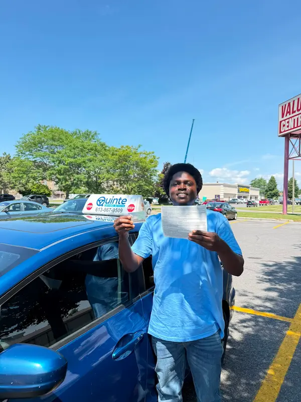 Smiling man wearing a blue t-shirt holding his road test passing paper in front of a Quinte Driving School car.