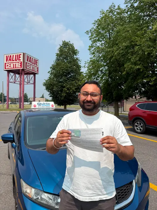 Smiling man wearing a white t-shirt holding his road test passing paper and his driving license in front of a Quinte Driving School car.