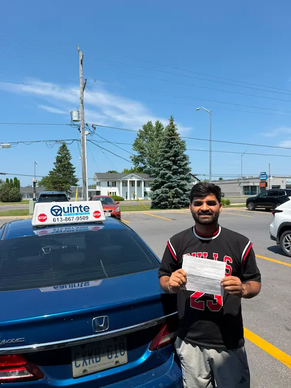 Smiling man wearing a black t-shirt holding his road test passing paper in front of a Quinte Driving School car.